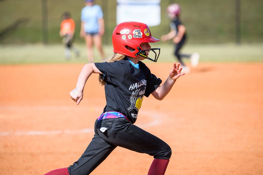 Youth softball player in black uniform running bases during game on dirt field.