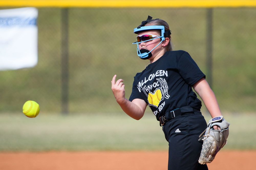Softball pitcher with protective face mask in mid-delivery motion.