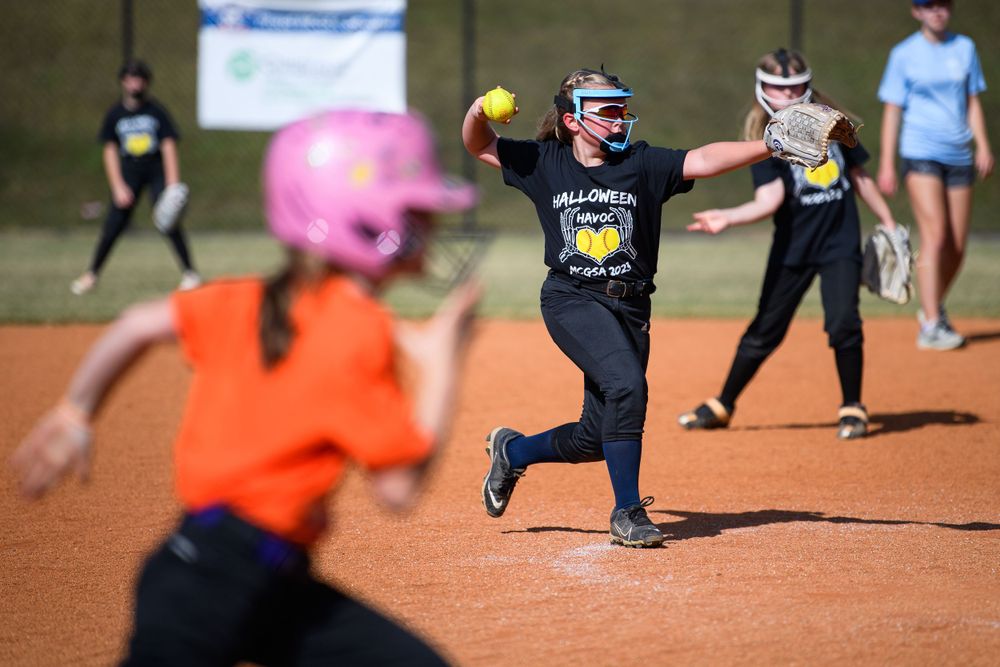 Softball pitcher in mid-delivery motion throwing yellow ball during game on dirt field.