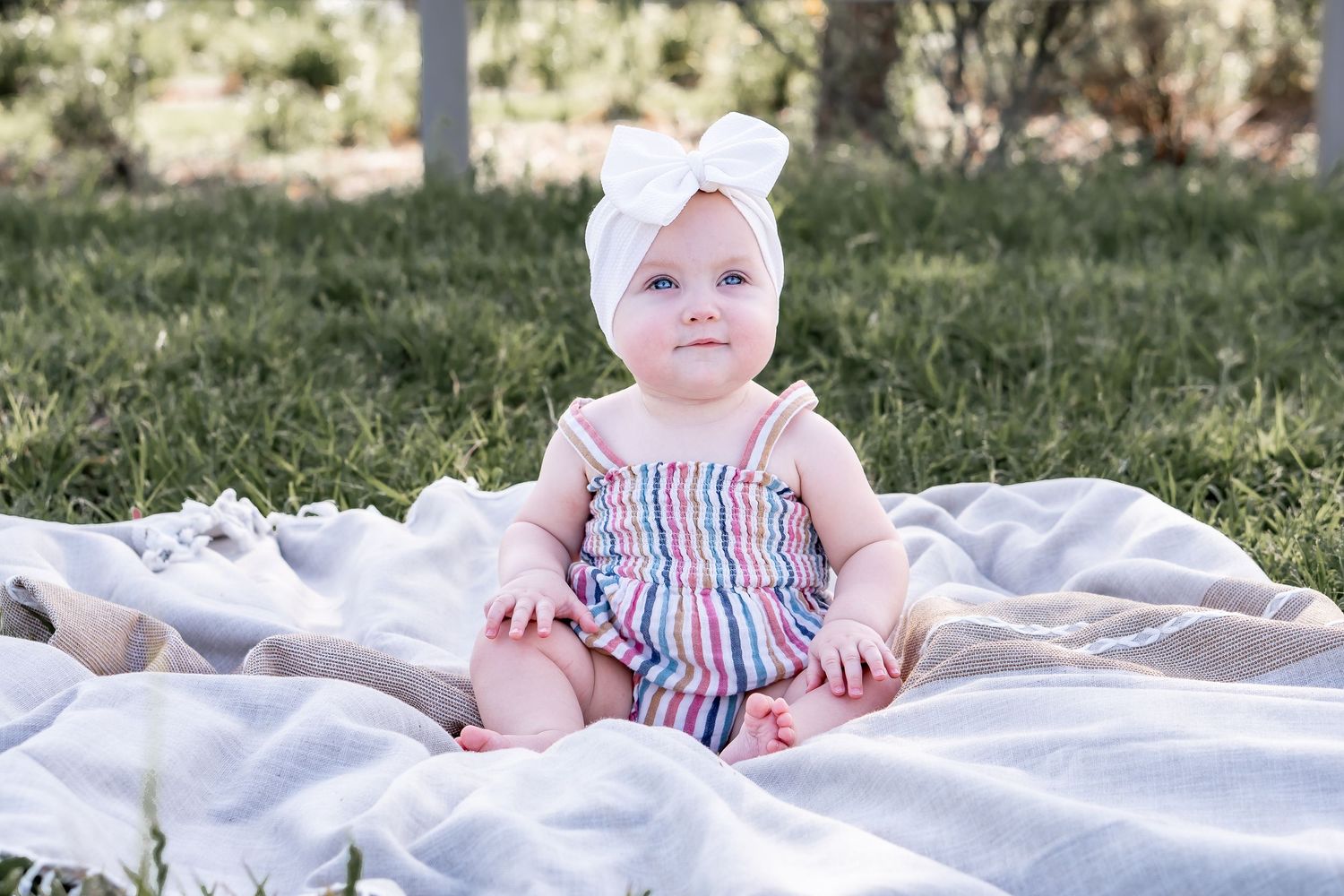 Baby in striped romper and white bow sits on white blanket in sunny garden setting.