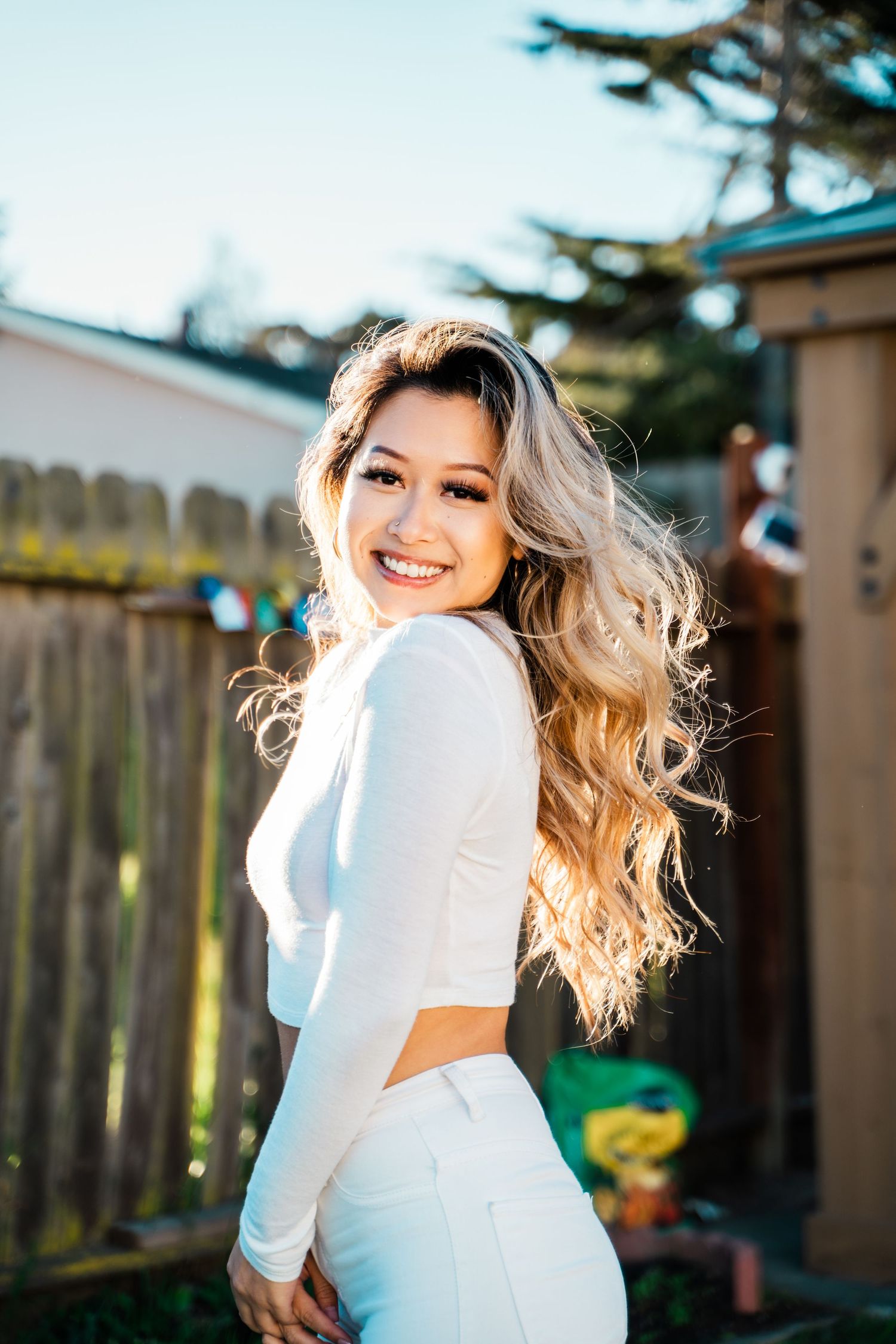 Outdoor portrait of a person with long wavy blonde hair wearing a white long-sleeved outfit against a backyard fence.