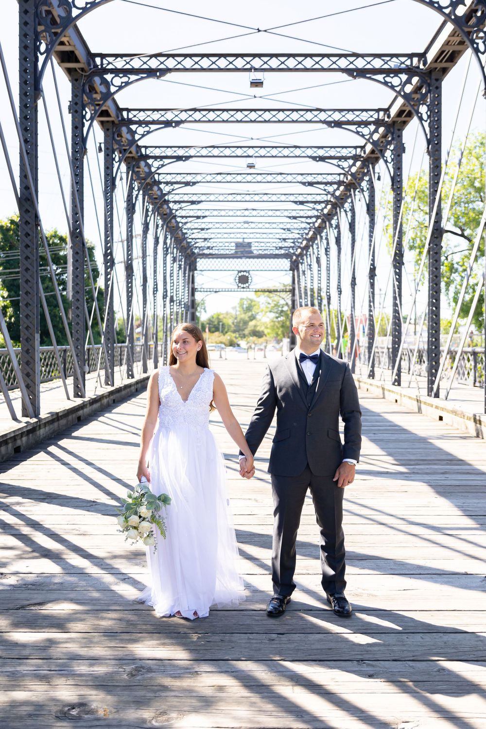 A couple in wedding attire walking together hand-in-hand on a historic metal bridge with geometric patterns and sunlight.