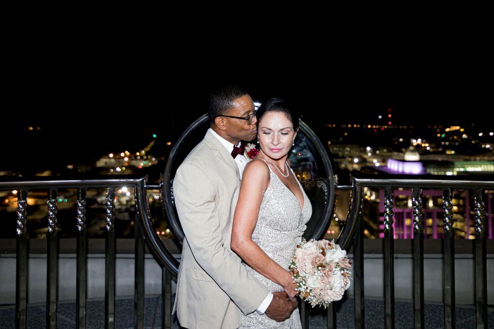 A newly married couple shares a romantic nighttime moment on a balcony overlooking twinkling city lights.
