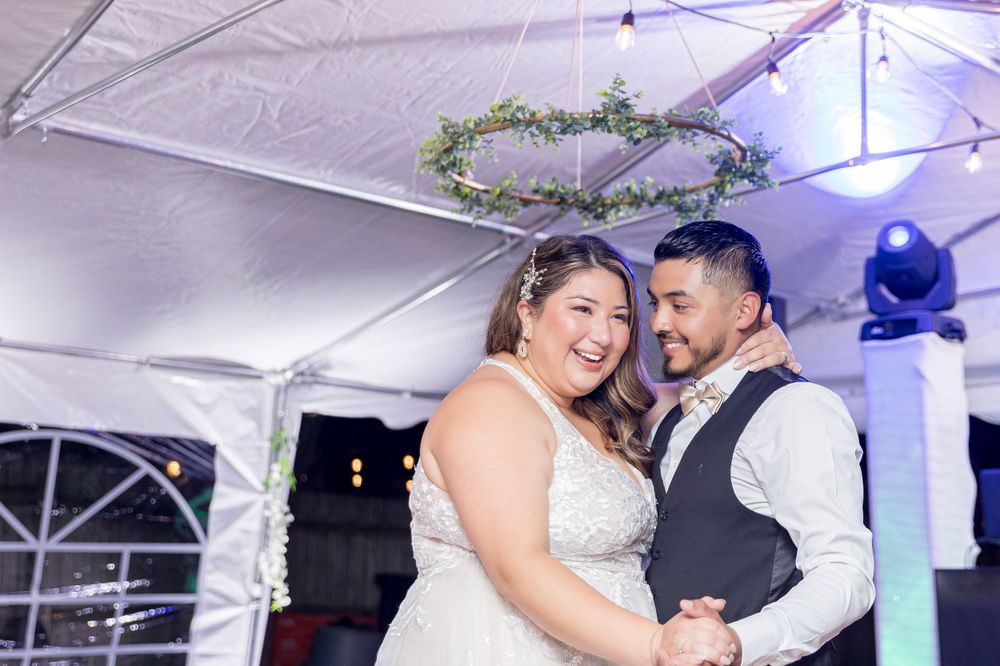 A couple shares a joyful dance moment under a decorated tent with a greenery hoop and purple lighting above.