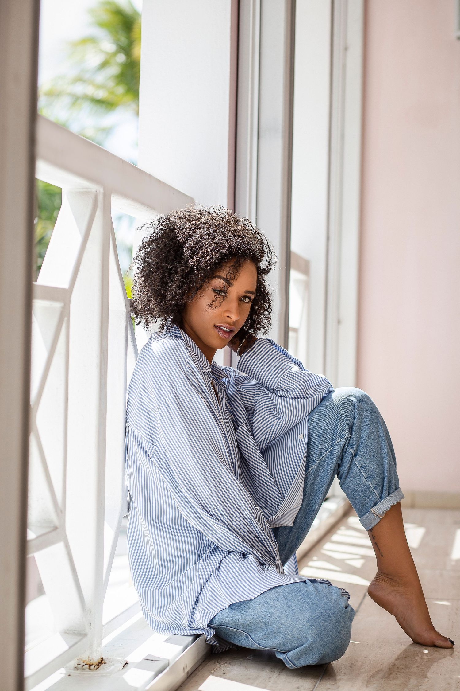 Woman with curly hair in striped shirt and jeans sits relaxed by a sunny window.