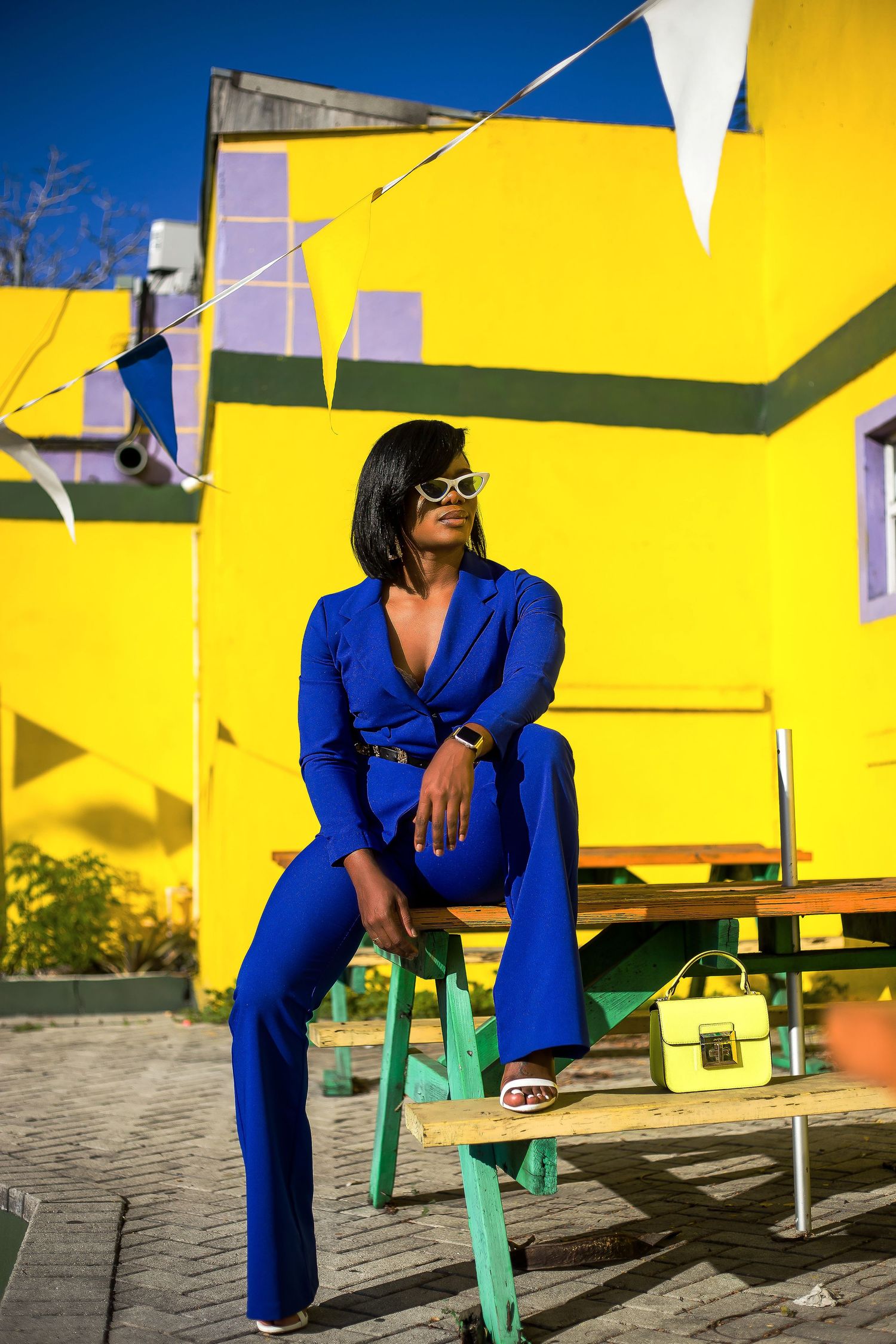 Woman in royal blue suit sits on bench before vibrant yellow building with festive bunting.