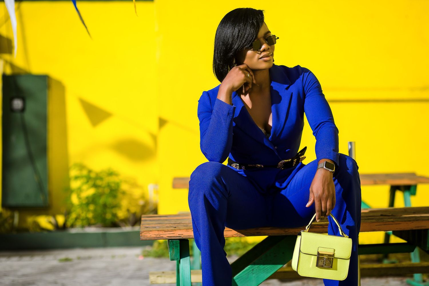 Fashionable woman in cobalt blue suit sits on wooden bench holding yellow handbag outdoors.