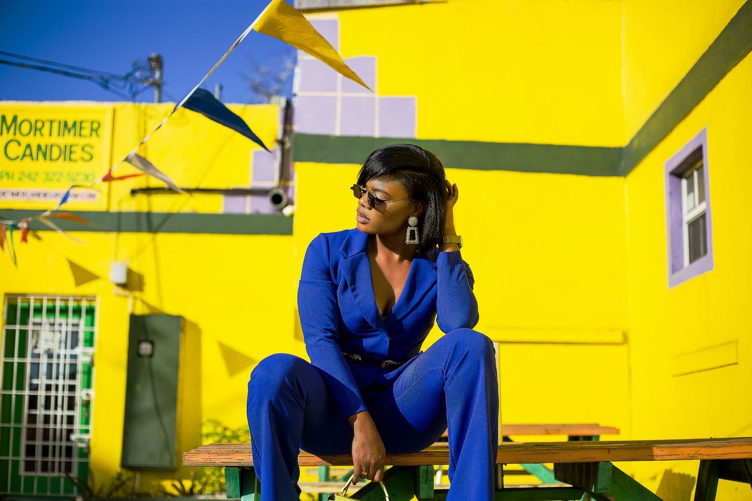 Stylish woman in blue jumpsuit poses at Mortimer Candies yellow storefront wearing sunglasses.
