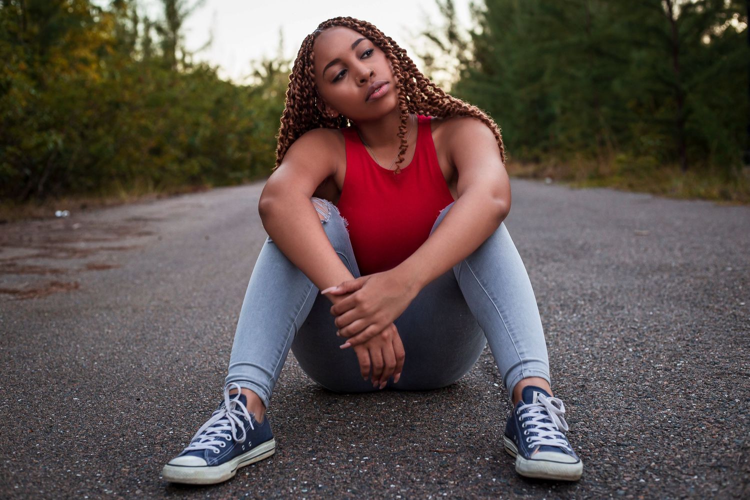 Young woman with braids sits on road in red top, jeans, and blue sneakers surrounded by trees.