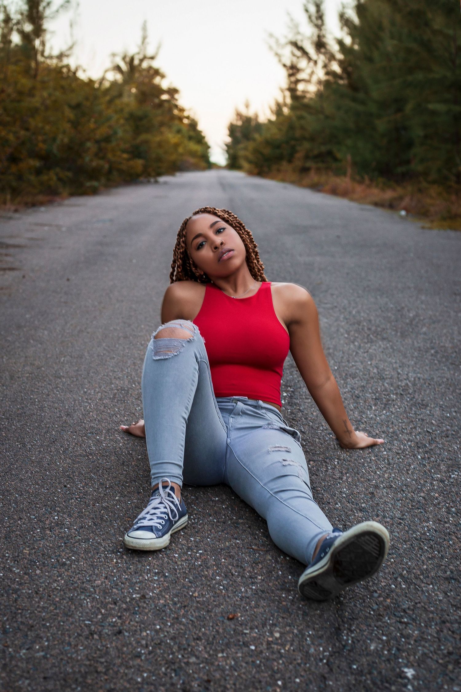 Young woman in red top and ripped jeans sits relaxed on an empty tree-lined road.