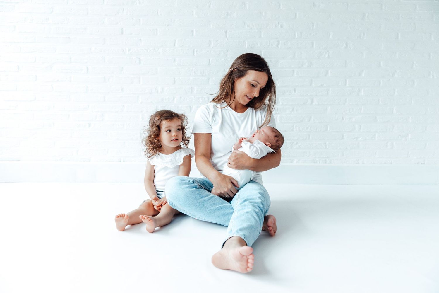 Mother in casual clothes sits on white floor with two children for family portrait session.
