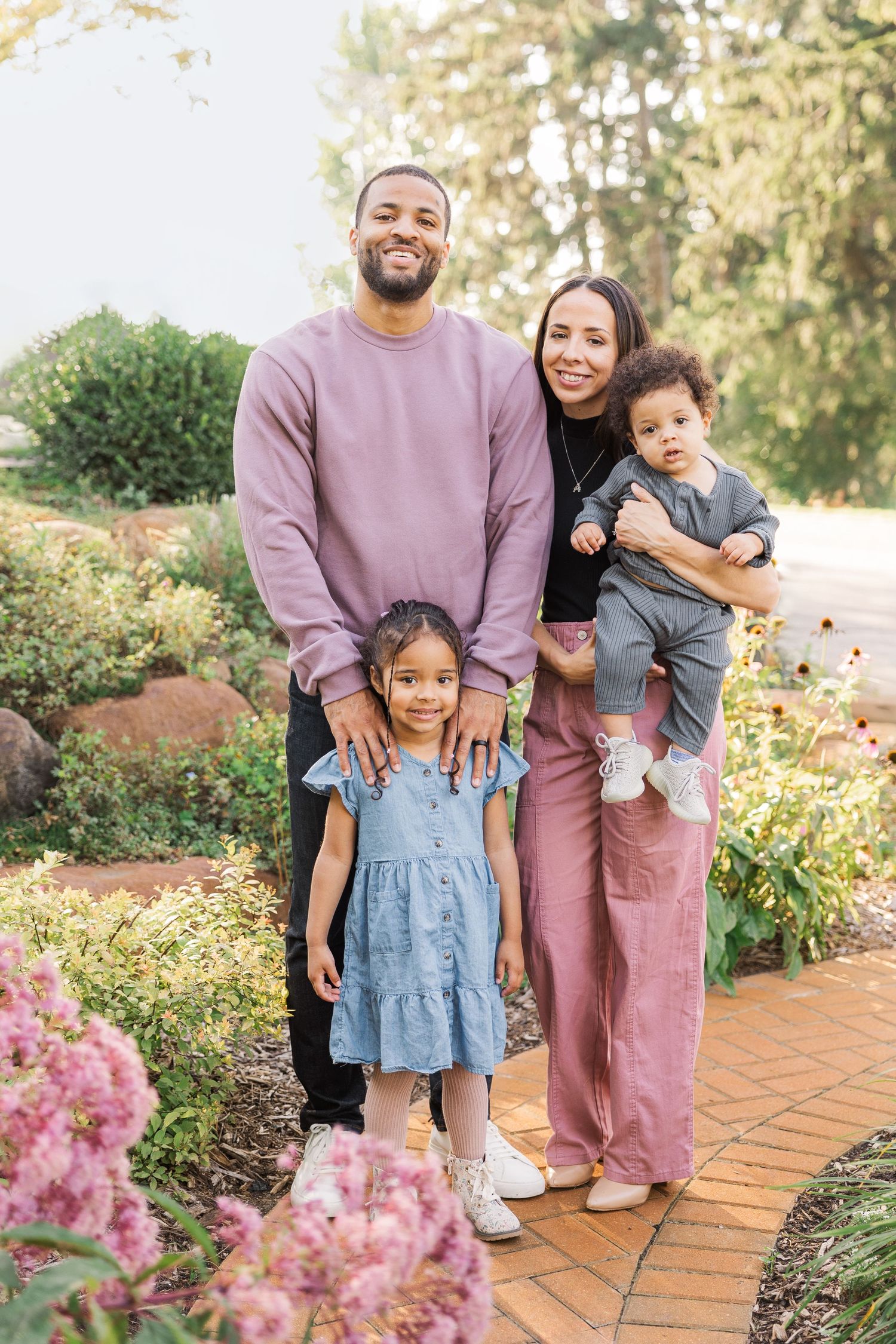 Family poses together on a brick path surrounded by blooming pink flowers in a garden. Novi Northville Portrait Photographer