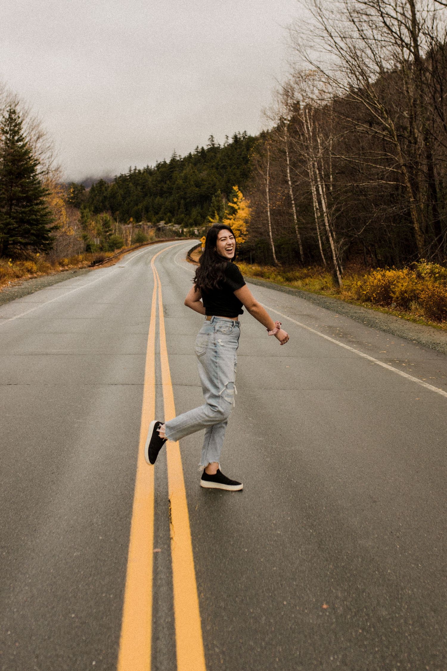 Jenna is standing in the middle of a road laughing as she spins around surrounded by trees in New Hampshire.