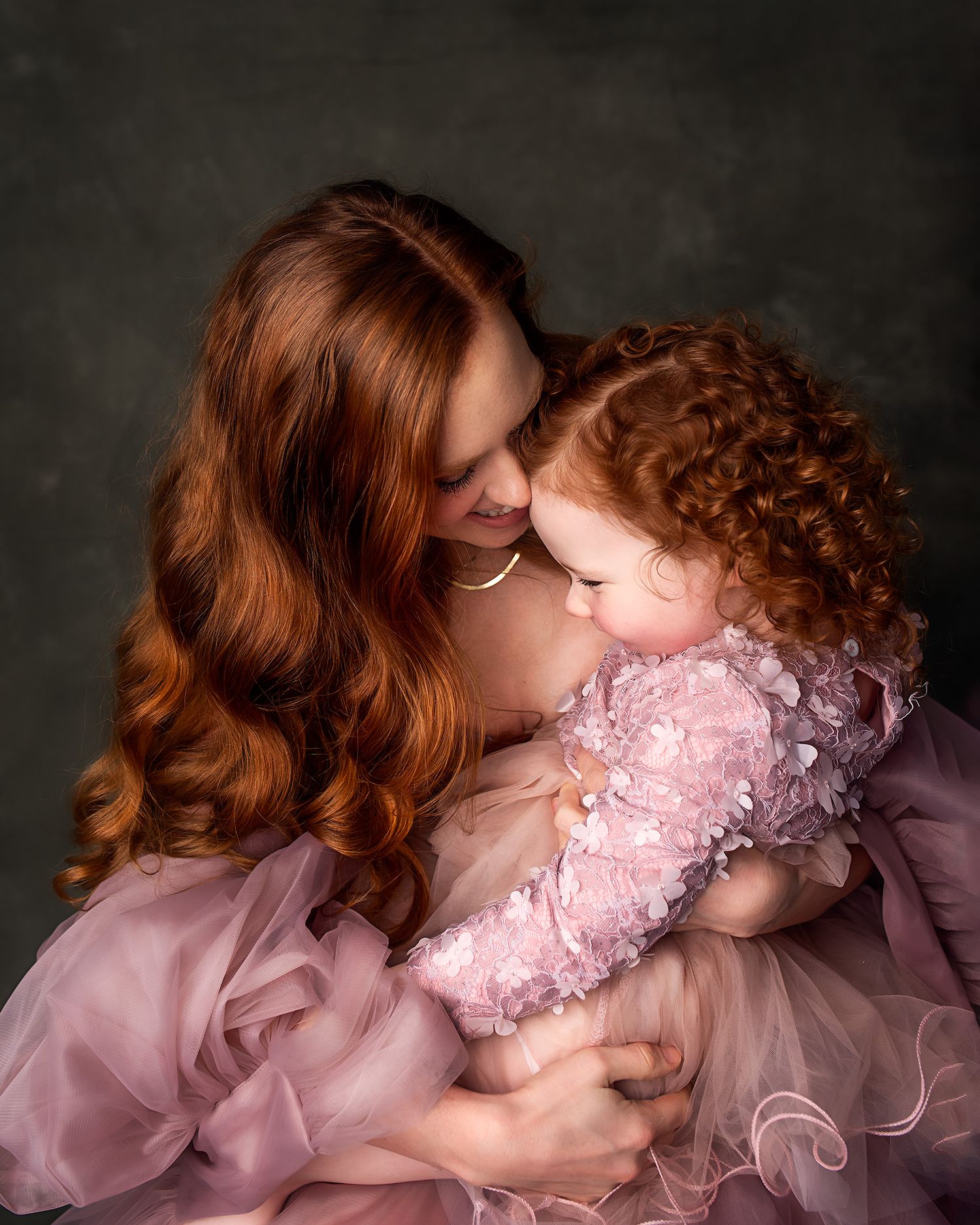 A mother with long red hair holds her curly-haired toddler in matching pink dresses in a studio portrait.