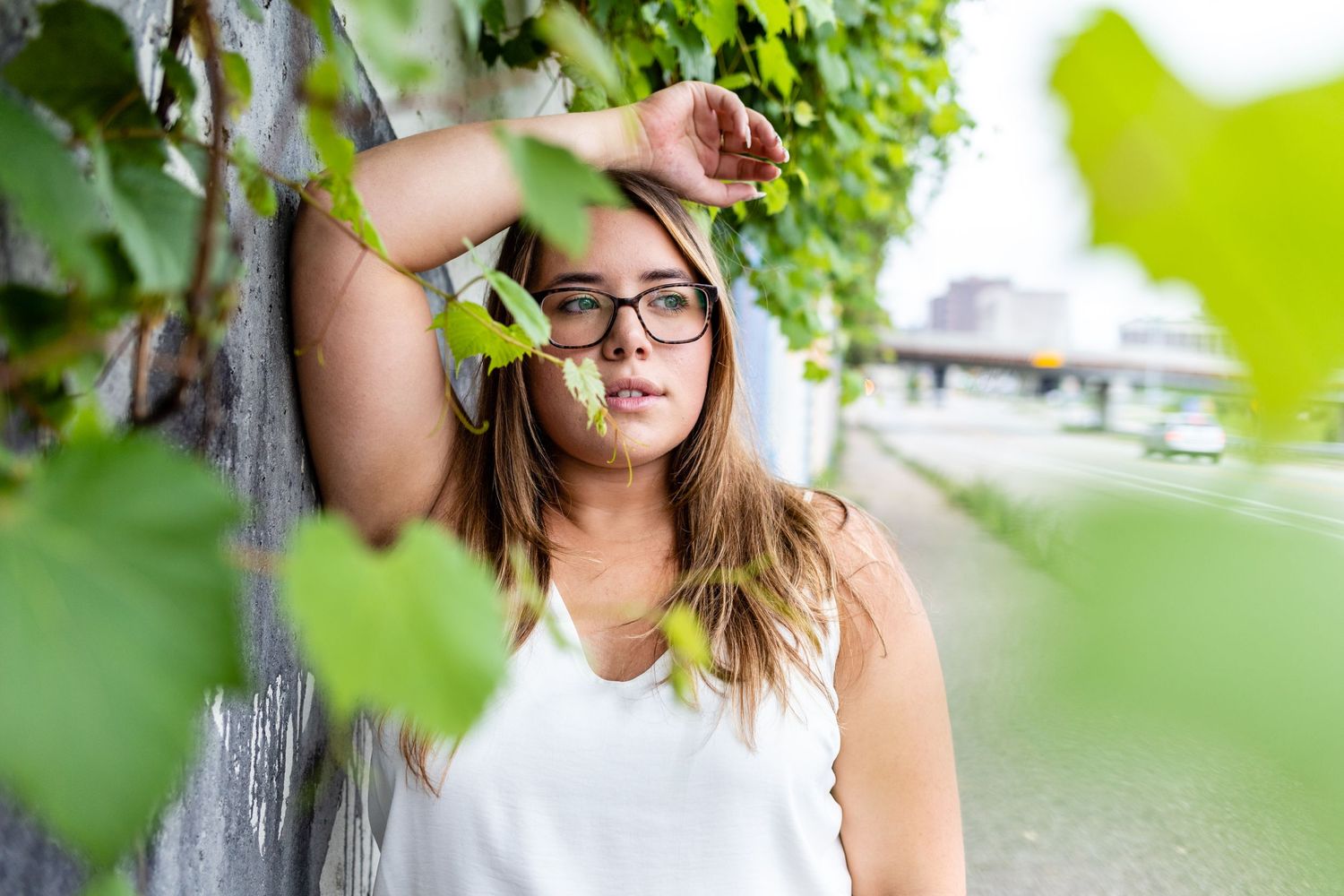 Young woman with glasses leaning against a wall surrounded by green ivy leaves.