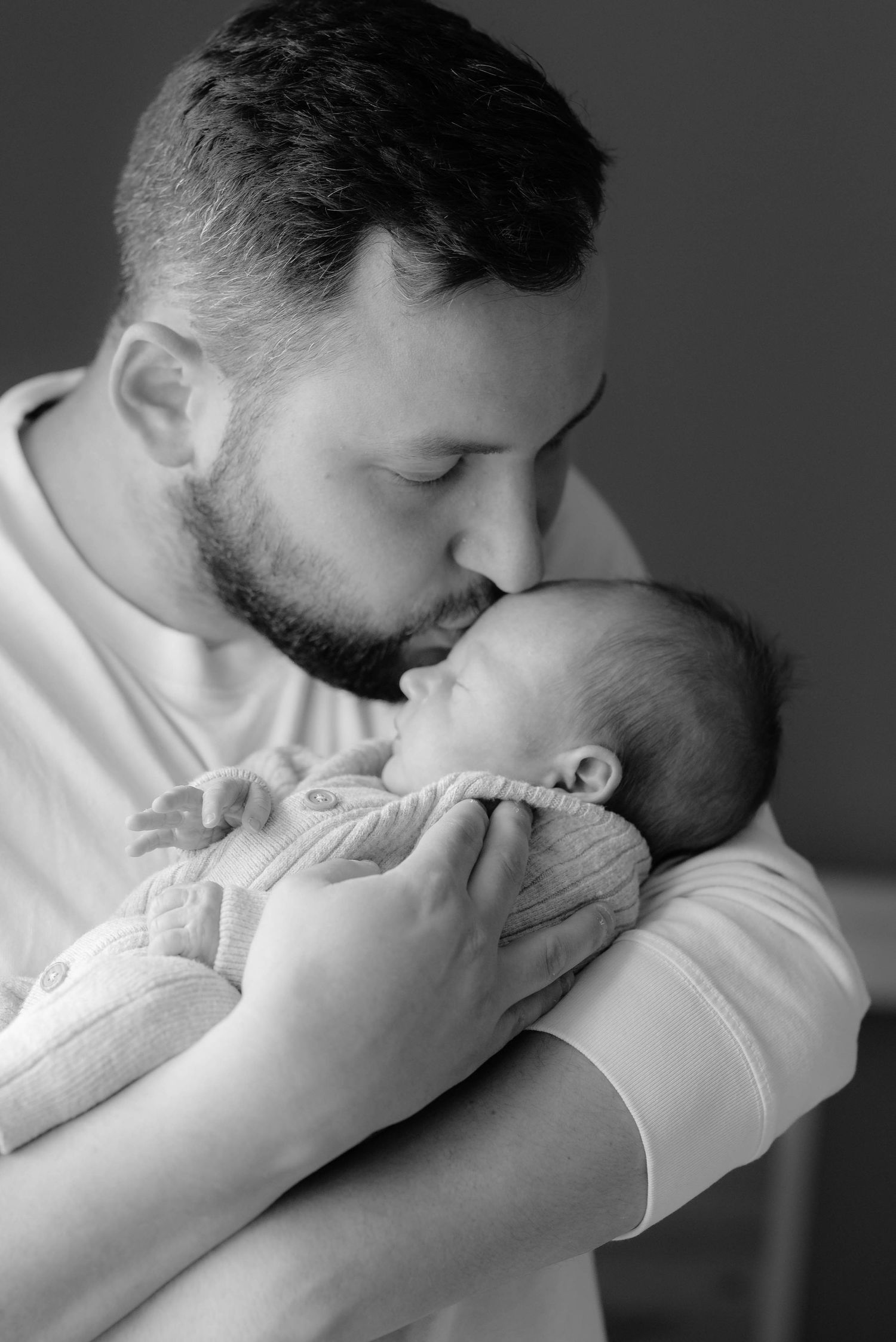 A tender black and white portrait of a parent gently kissing a newborn baby wrapped in light fabric.