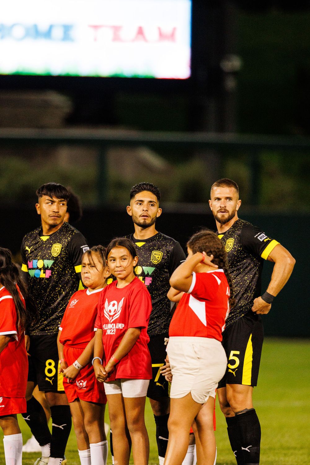 Soccer players pose with young fans in red jerseys on field.