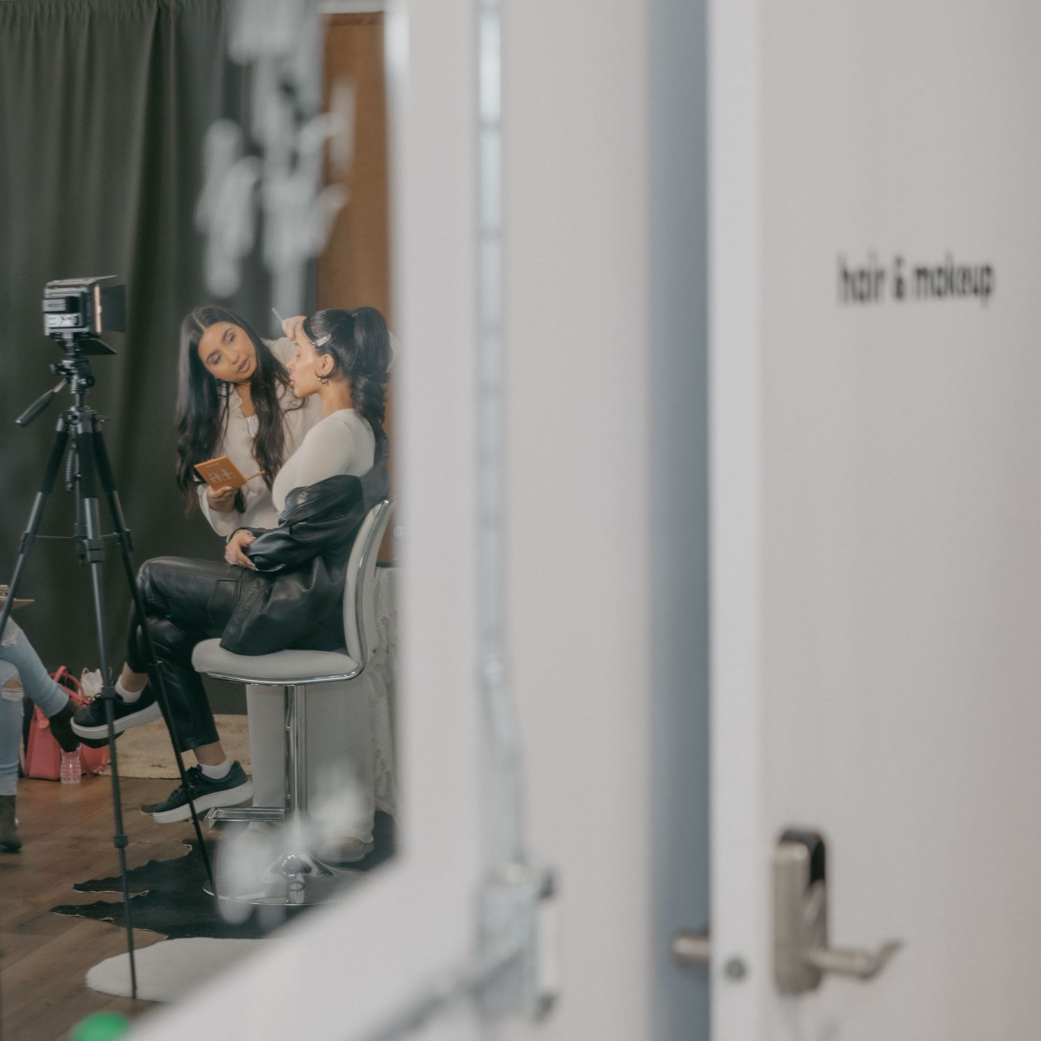 A person sits in a chair next to a camera tripod in a minimalist studio setting.