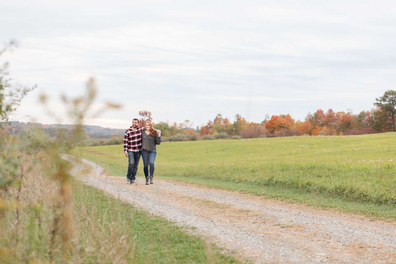 A couple walks down a gravel path surrounded by autumn trees and rolling hills in the countryside.