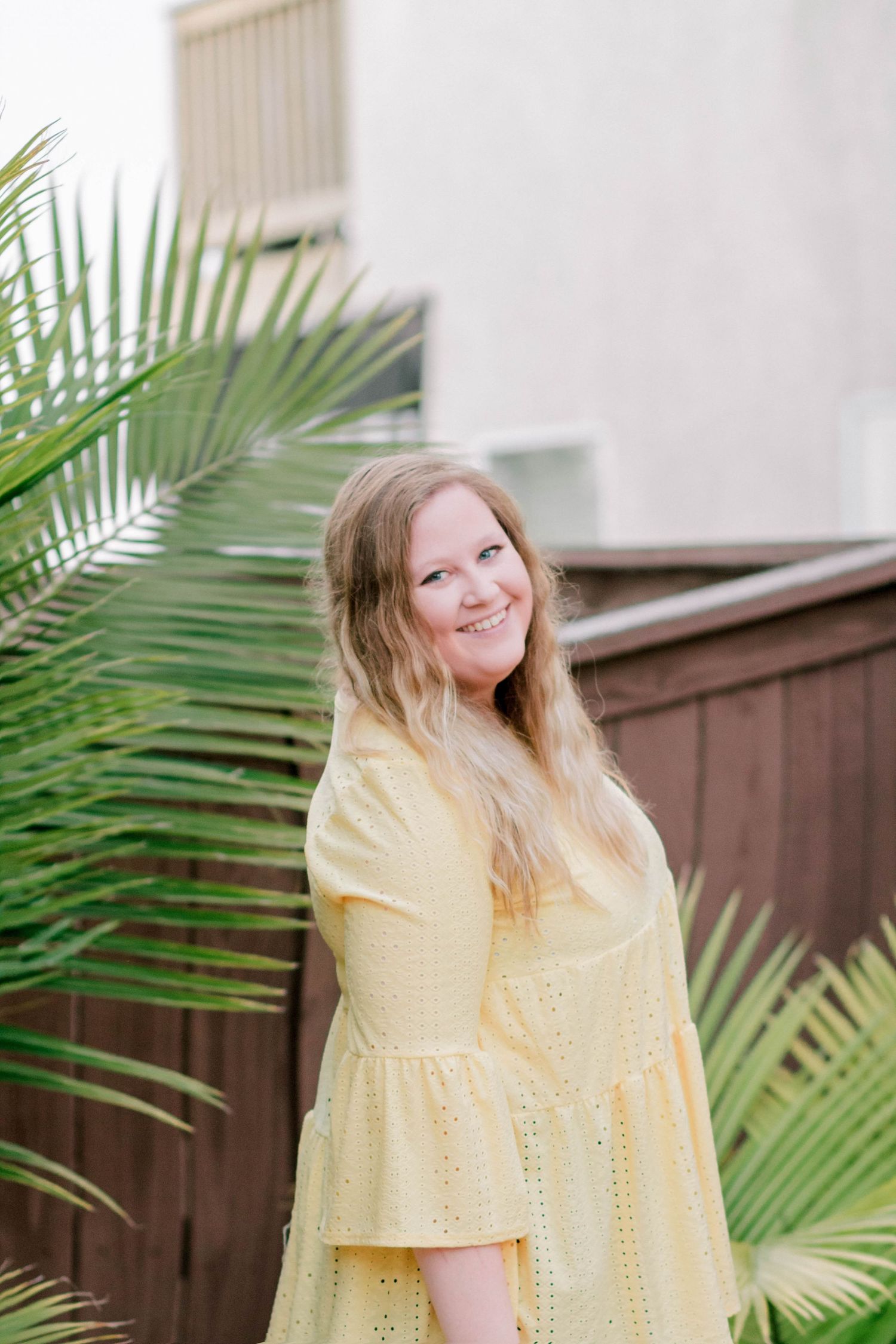 Smiling young woman in yellow eyelet dress posing near tropical palm leaves in outdoor setting.