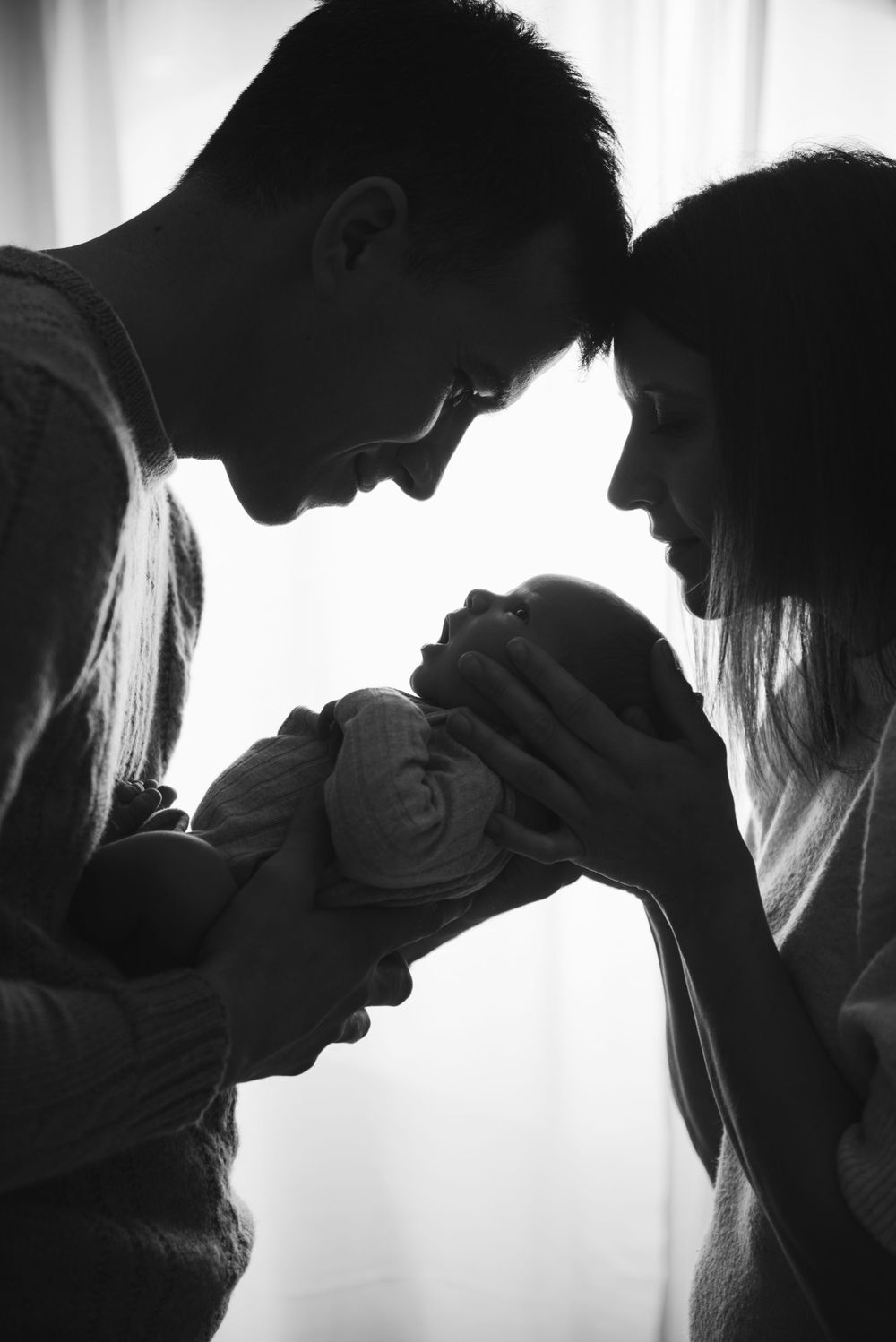 Silhouetted figures share an intimate moment with newborn against window light.