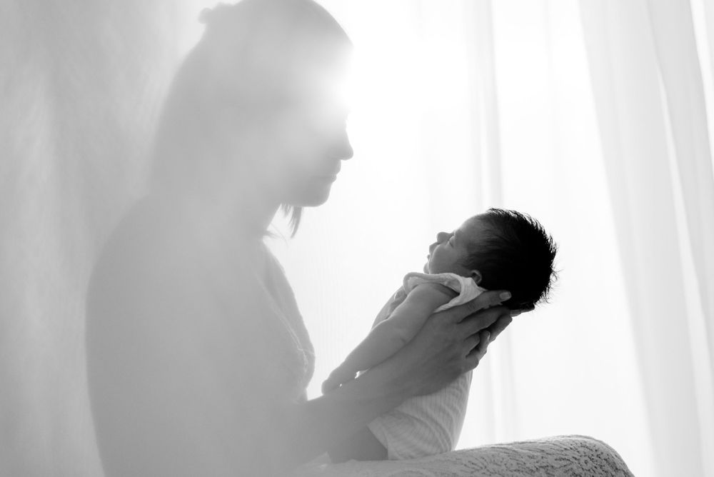 Silhouette of person holding baby against bright window background.