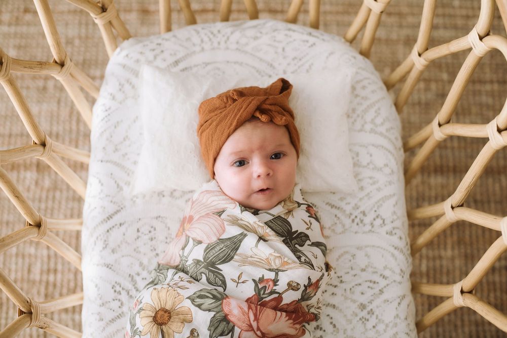 Baby wearing floral swaddle and orange headwrap lying in white textured bassinet.