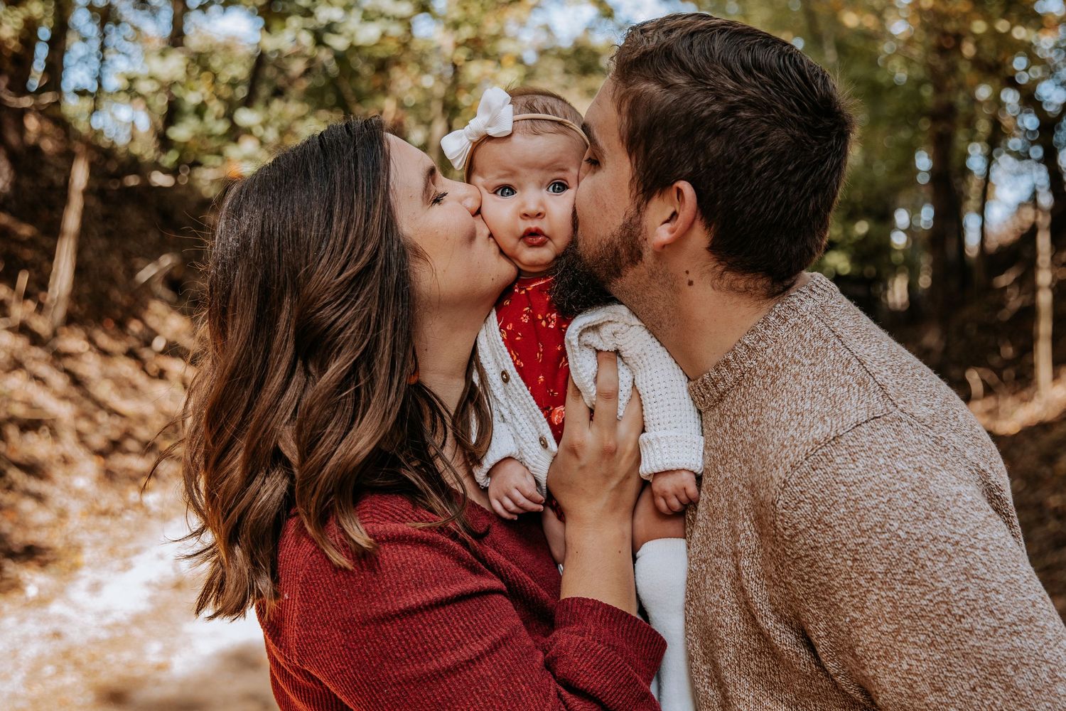 A loving family moment captured outdoors as parents kiss their baby's cheeks.