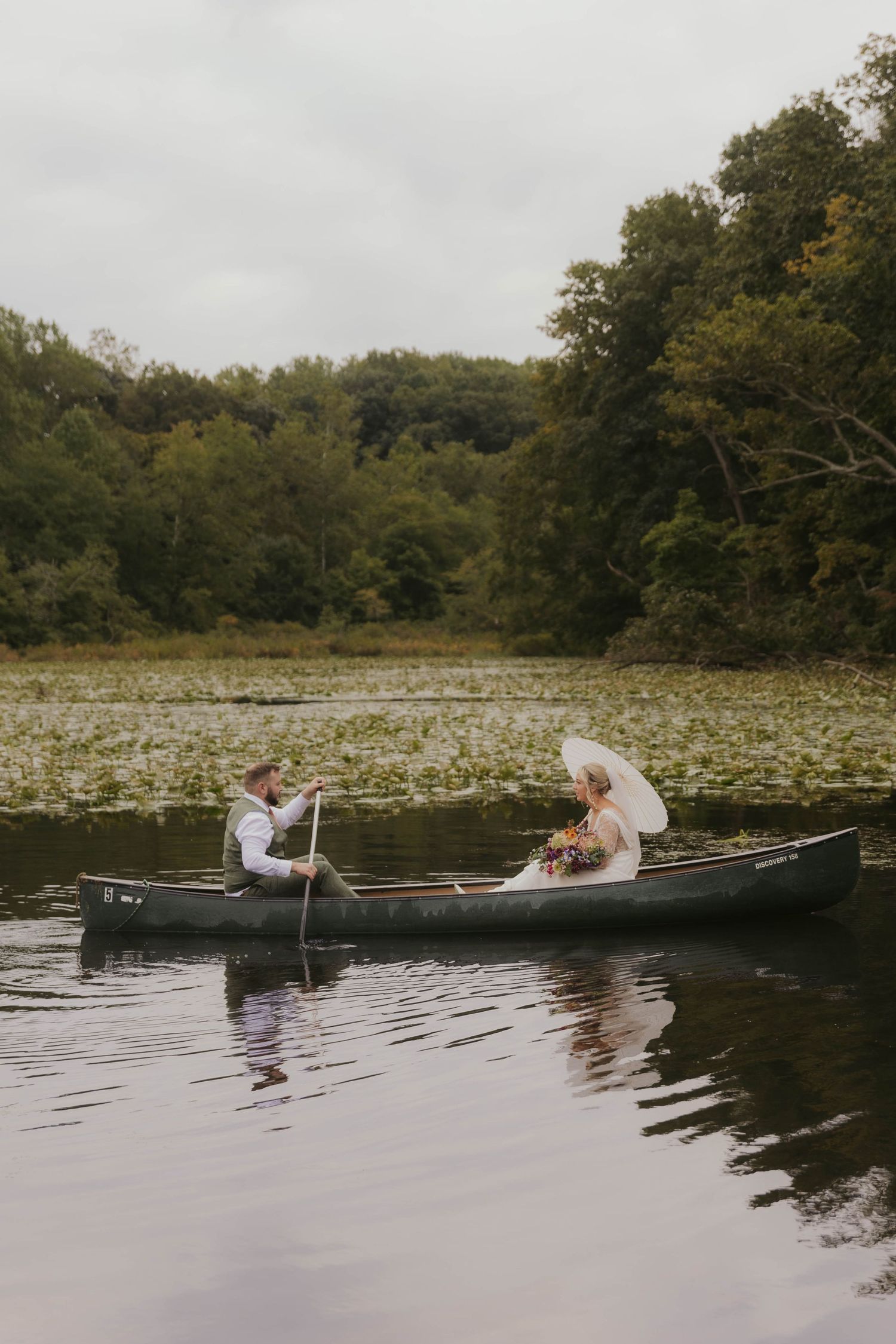 Wedding couple paddling in canoes across a serene lily pad covered lake.