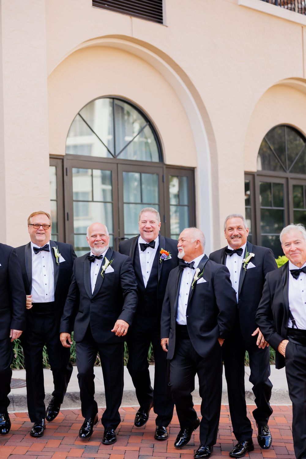 Wedding party members in tuxedos share a candid moment outside a Mediterranean-style building.
