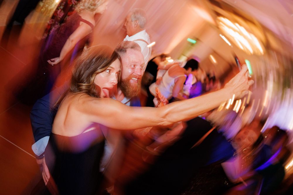 Blurred motion shot of wedding guests dancing energetically at a reception with purple lighting.
