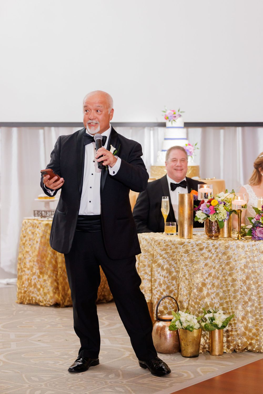 Speaker in tuxedo delivers toast at wedding reception with gold-decorated head table.
