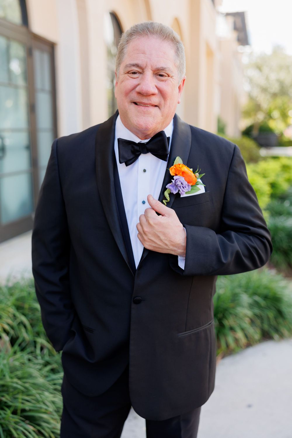 A gentleman in a classic black tuxedo with an orange boutonniere poses outside an elegant venue.
