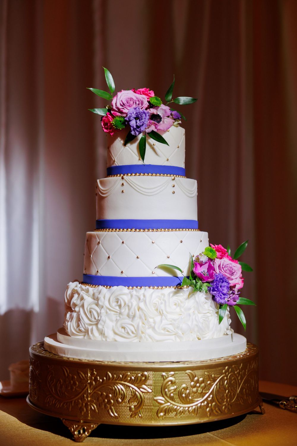Elegant four-tier white wedding cake with blue ribbon trim and fresh pink and purple floral decorations.