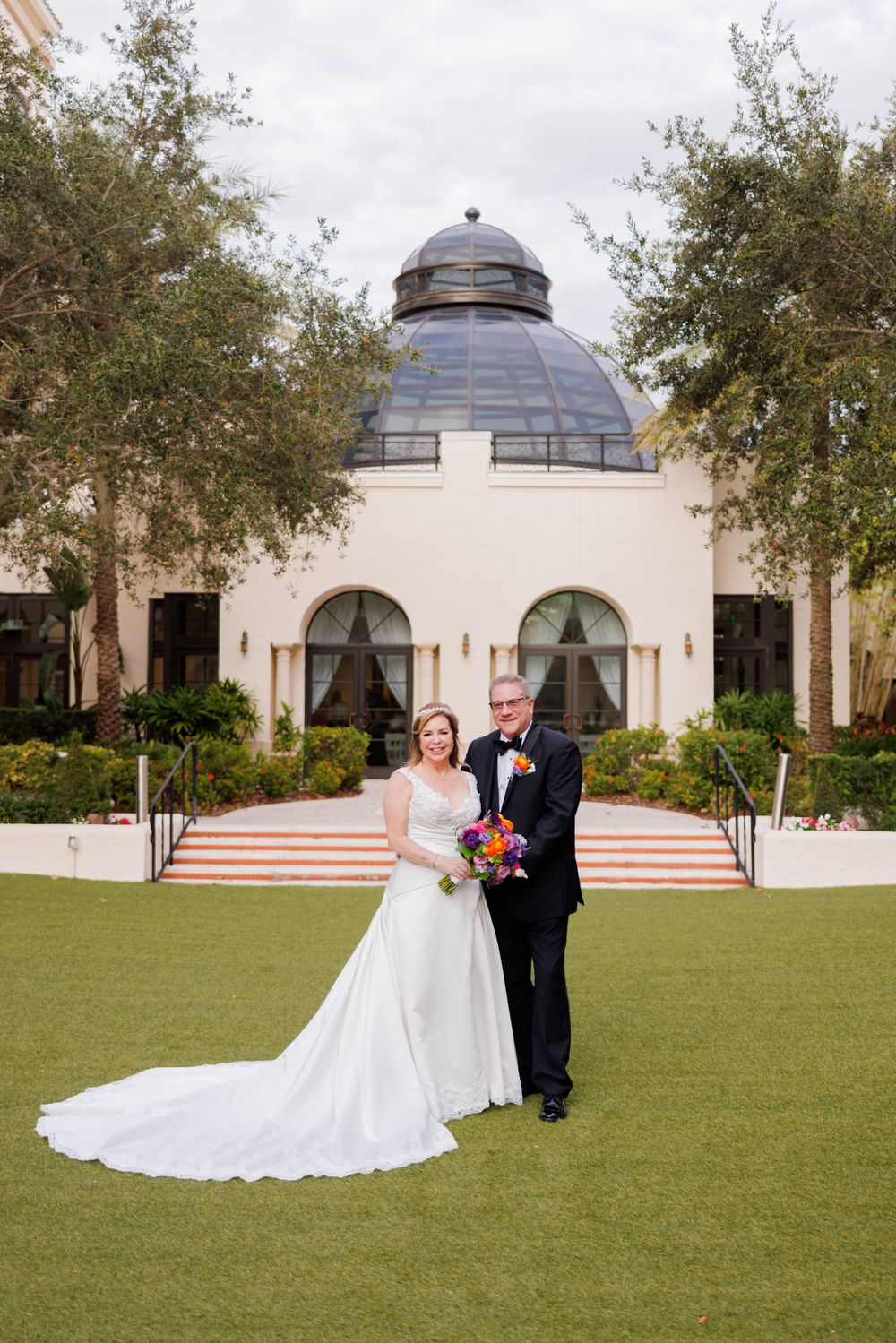 A wedding photo in front of a domed building with white facade and grand entrance steps.