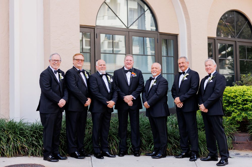 Groomsmen in black tuxedos pose for a formal group photo in front of an arched building entrance.