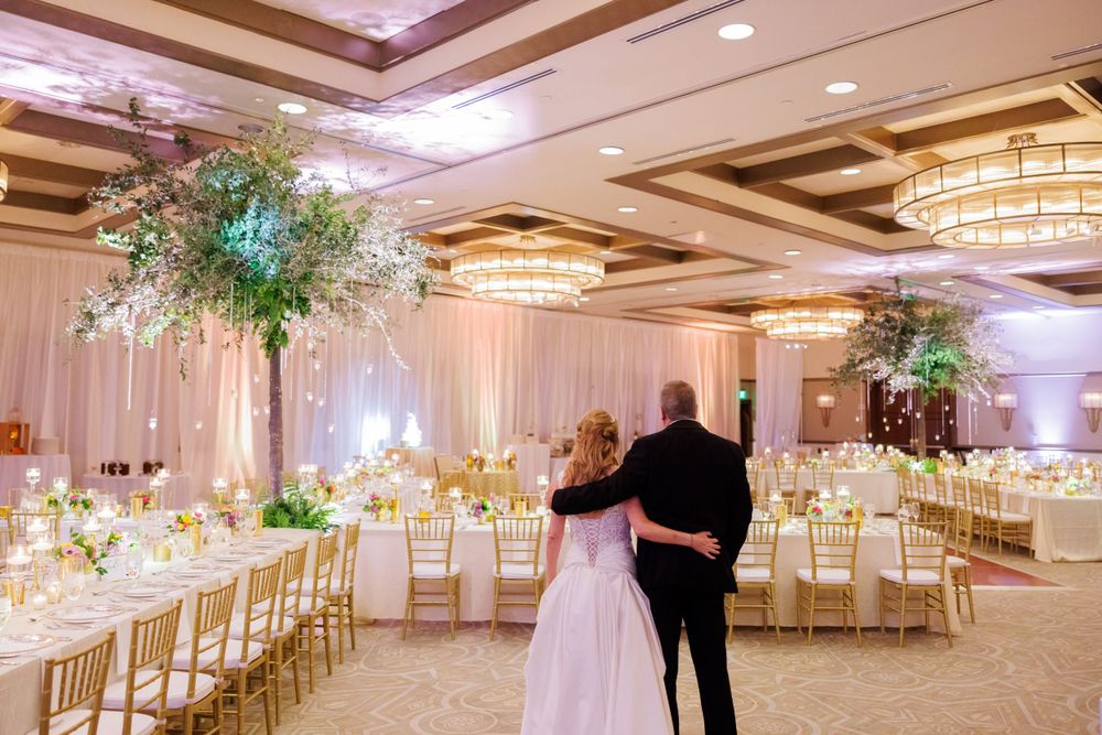 Elegant ballroom wedding reception with white draping, gold chairs and tall floral centerpieces illuminated by crystal chandeliers.