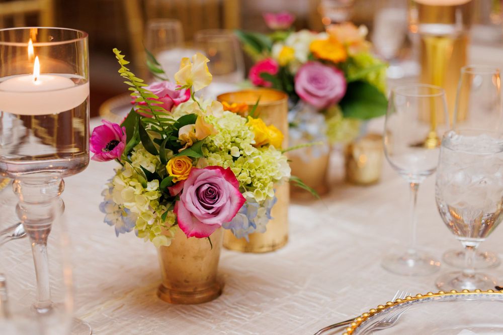 Colorful floral centerpiece with pink roses and yellow blooms in gold vase on elegant place setting.