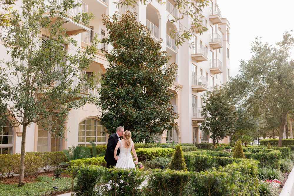 A couple poses in front of an elegant beige building with arched windows and manicured garden landscaping.
