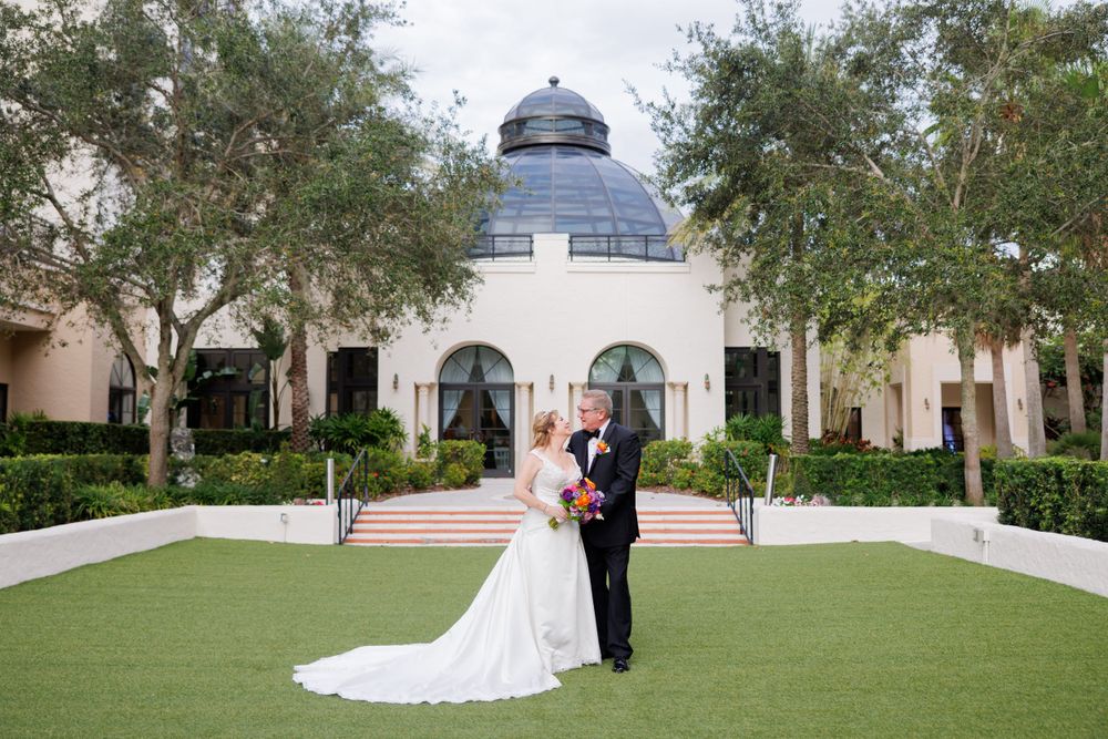 A couple stands on manicured lawn in front of a beautiful domed architectural structure.