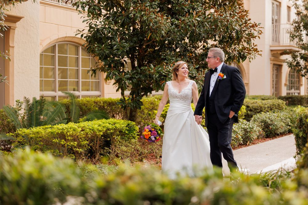 A couple walks along a garden path beside a Mediterranean-style building with ornate architecture.