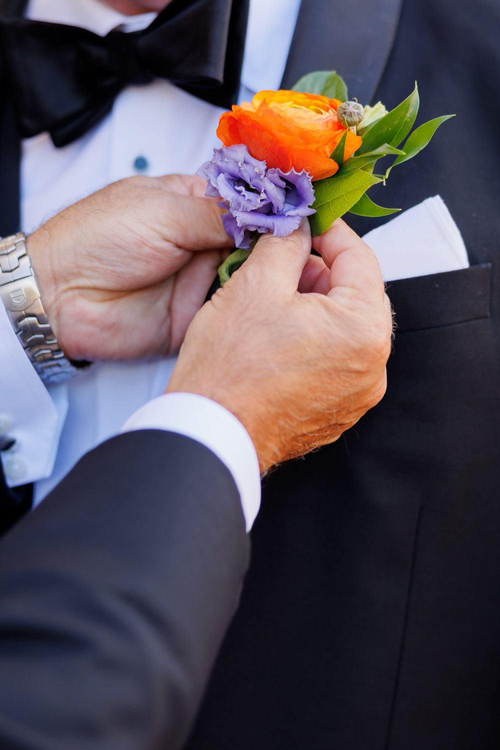 Hands adjusting a colorful boutonniere with orange and purple flowers on a formal tuxedo.