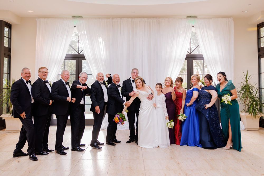 Large wedding party posed together in front of white draping with groomsmen and bridesmaids.