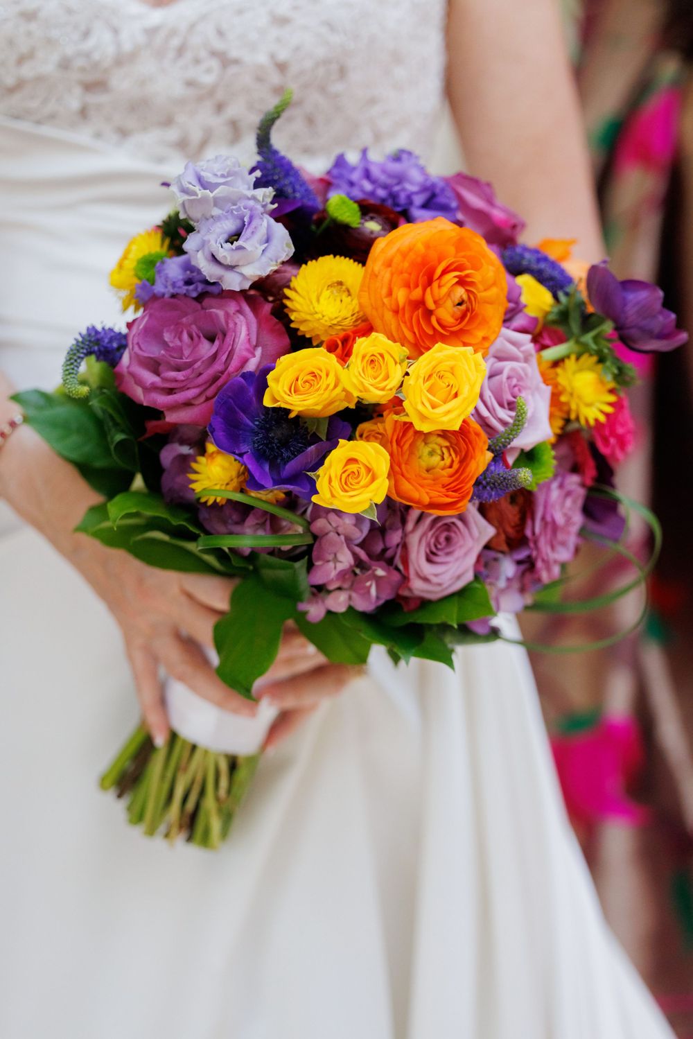 Vibrant wedding bouquet with purple, yellow, and orange roses against white dress fabric.