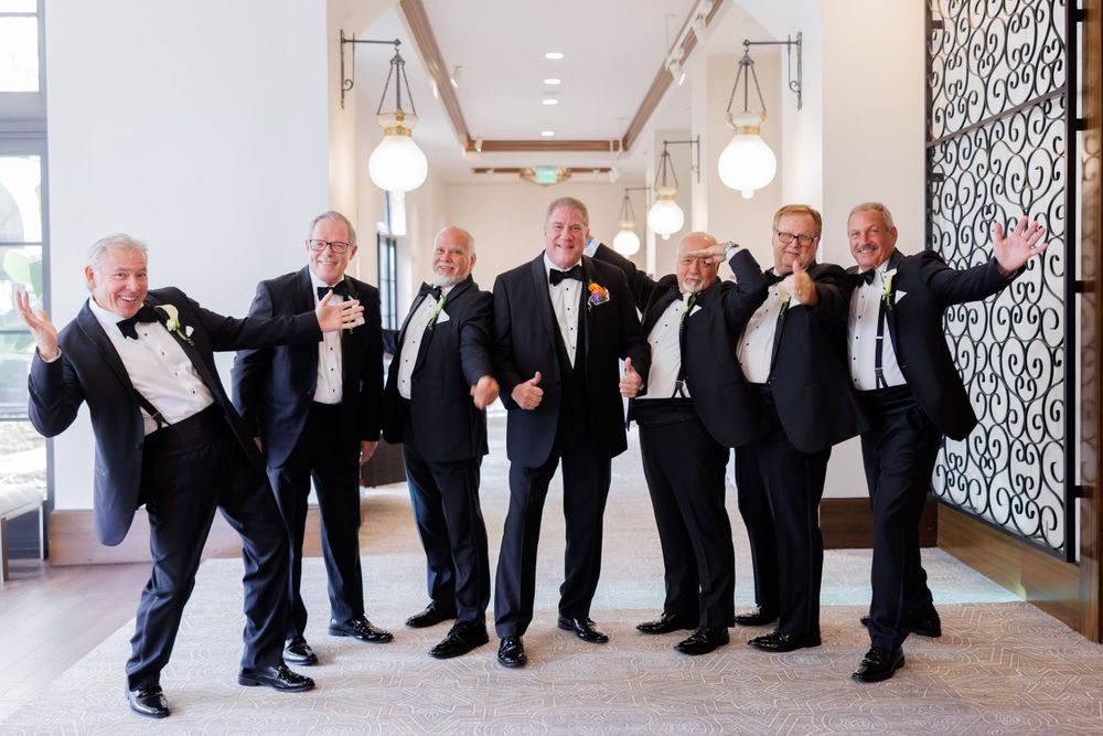 Groomsmen in tuxedos strike playful poses in an elegant hallway with pendant lighting.
