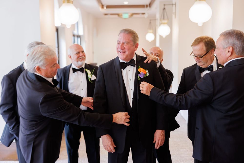 Groomsmen in classic black tuxedos striking playful poses in an elegant hallway.