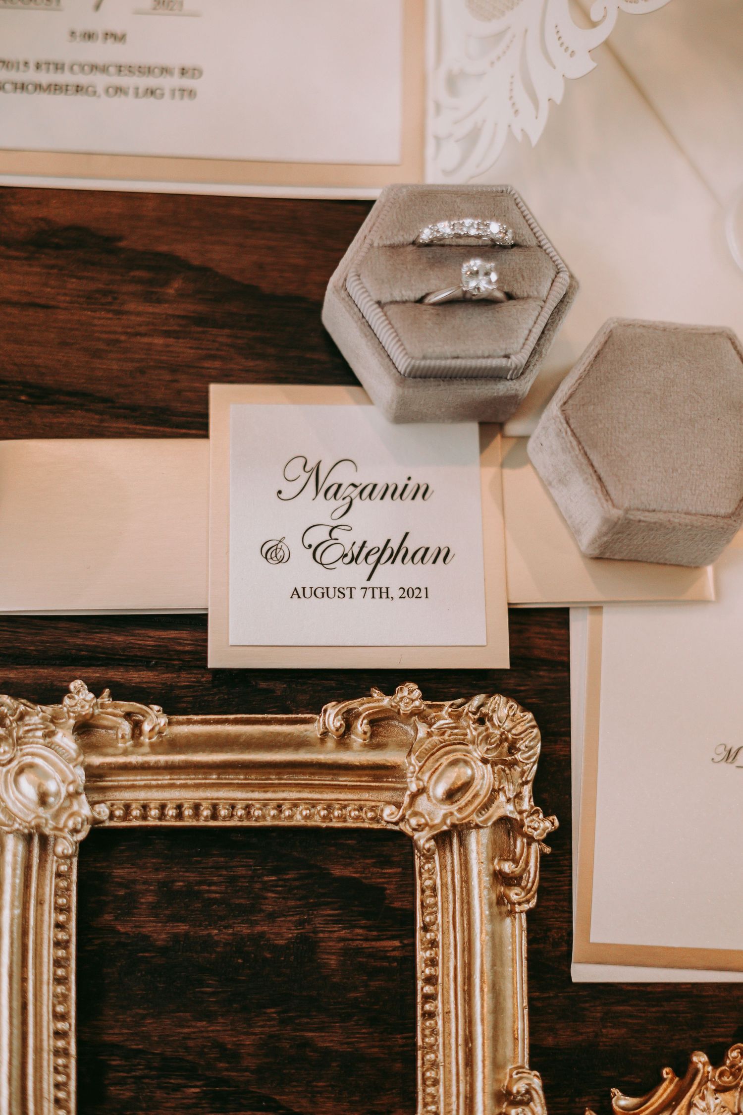 Wedding rings displayed in hexagonal boxes next to elegant name cards and ornate golden frame on dark wood surface.