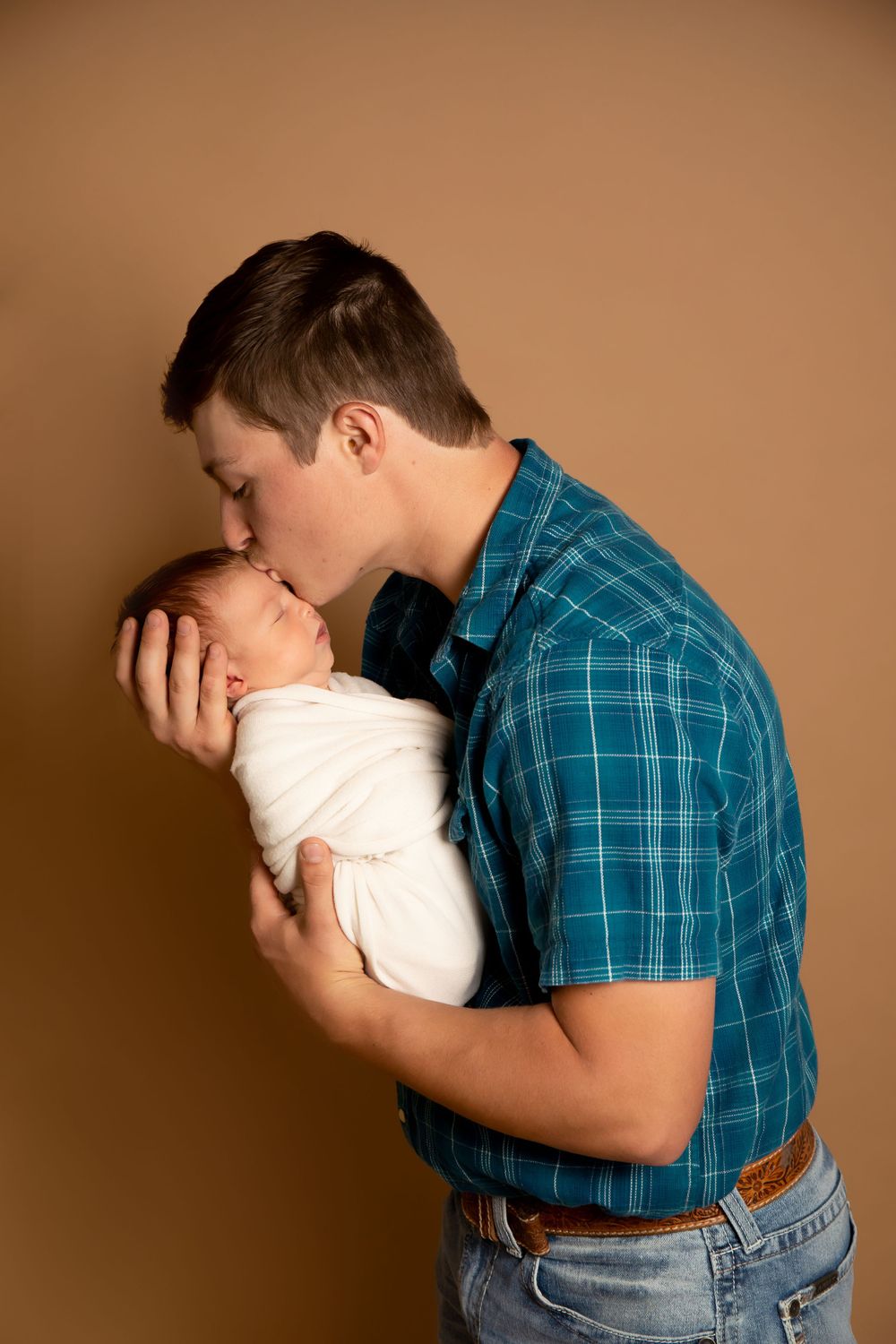 A tender moment between parent and newborn baby swaddled in white against a brown background.
