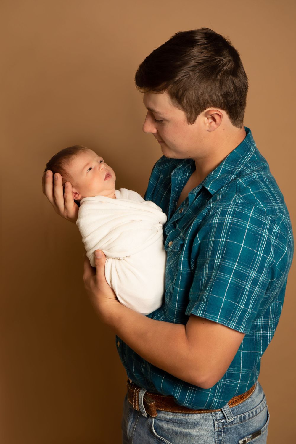 Tender moment between parent and swaddled newborn against brown backdrop.