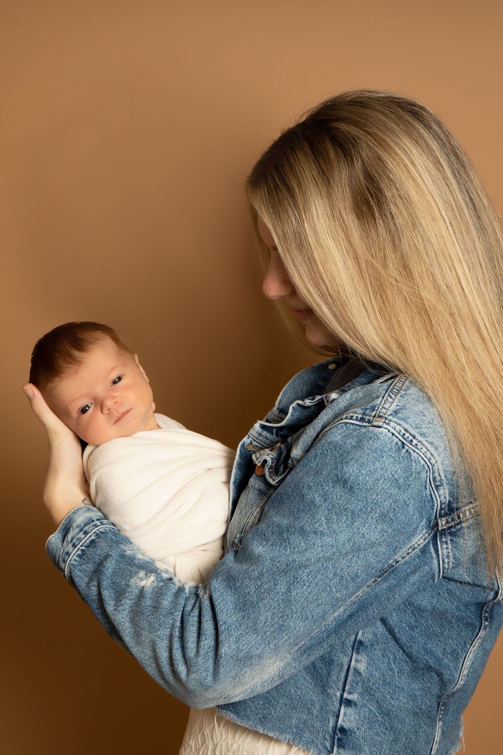 Parent in denim jacket holding swaddled baby against warm brown background.